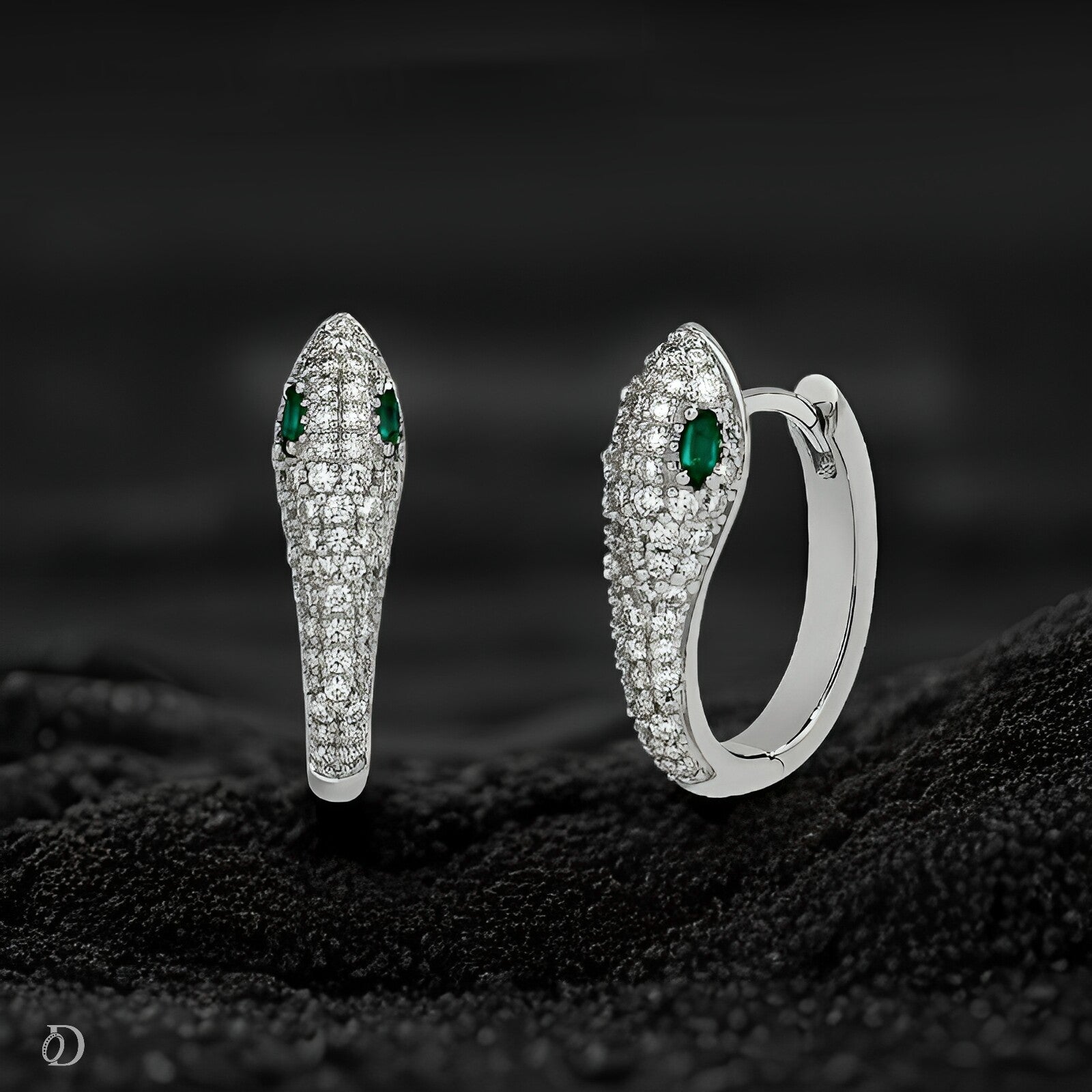 Silver snake-shaped earrings with green gemstones on a textured black surface