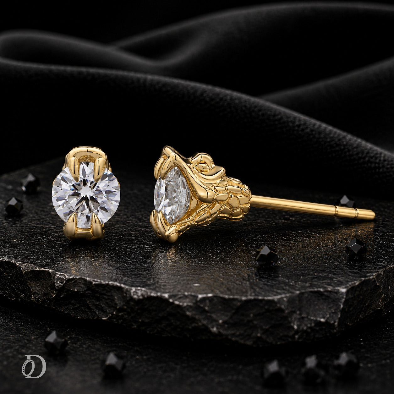 A pair of gold stud earrings with clear gemstones, displayed on a textured black stone slab with draped black fabric in the background.