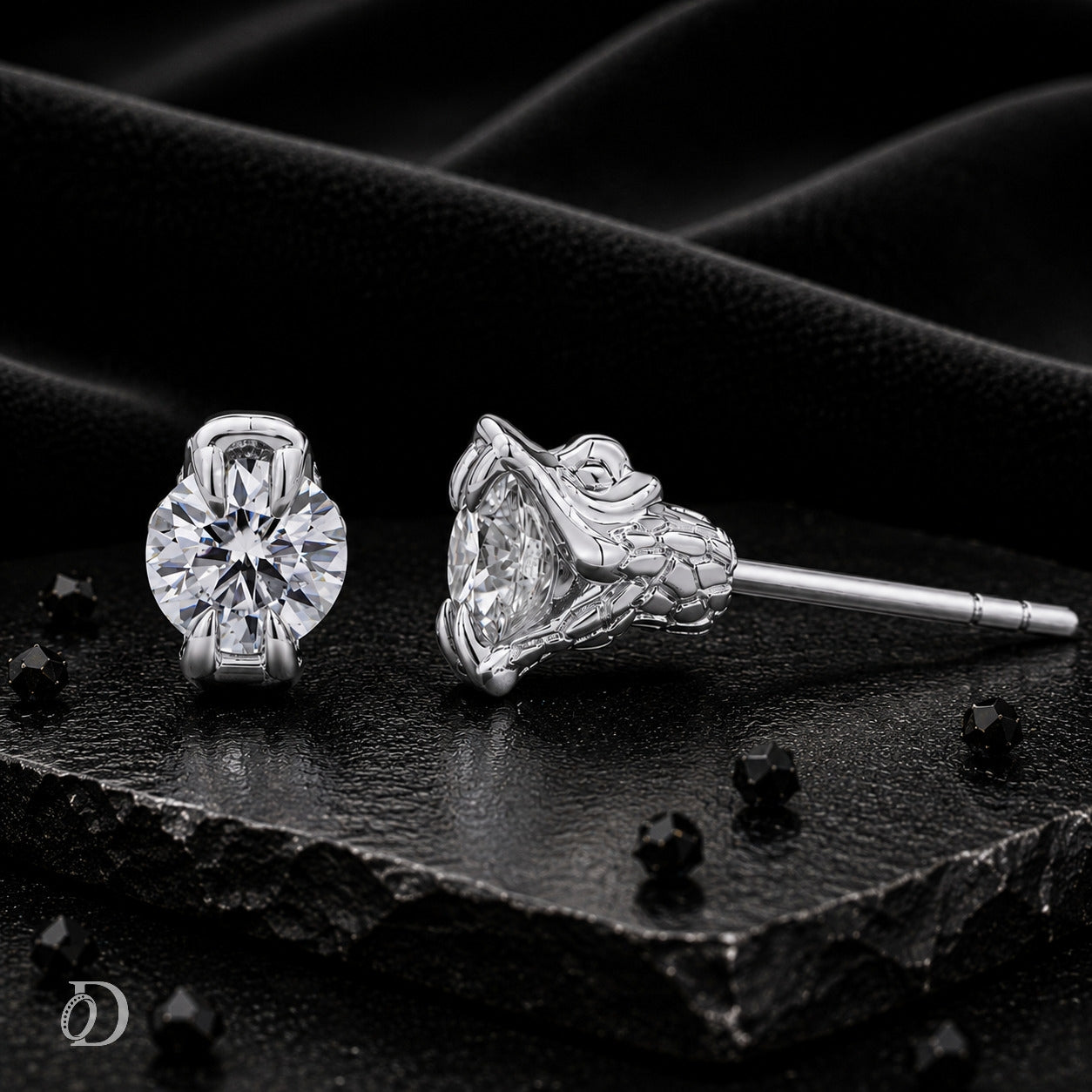 A pair of silver stud earrings with large clear gemstones, one shown from the front and the other from the side, displayed on a textured black surface with draped black fabric in the background.