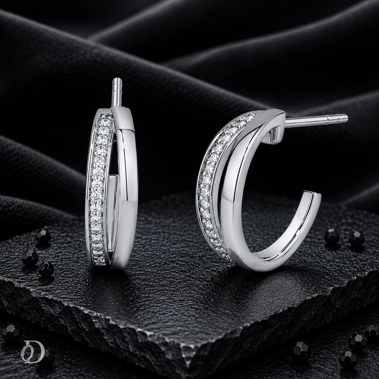 A pair of silver hoop earrings with clear pavé-set stones, displayed on a textured black stone surface with draped black fabric in the background.