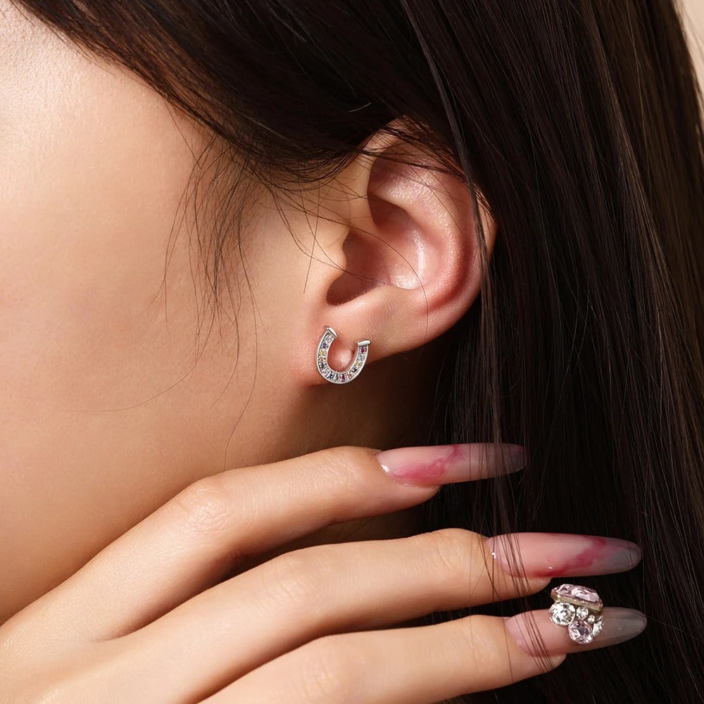 A close-up lifestyle shot of a model wearing a small silver crescent-shaped hoop earring adorned with clear crystals. The model has dark brown hair, fair skin, and manicured nails with a matching crystal ring on one finger.