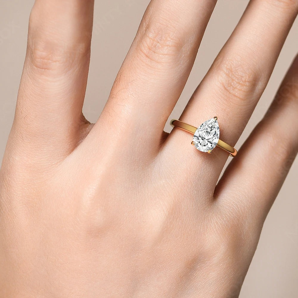 Hand wearing a gold ring with a pear-shaped diamond on a neutral background