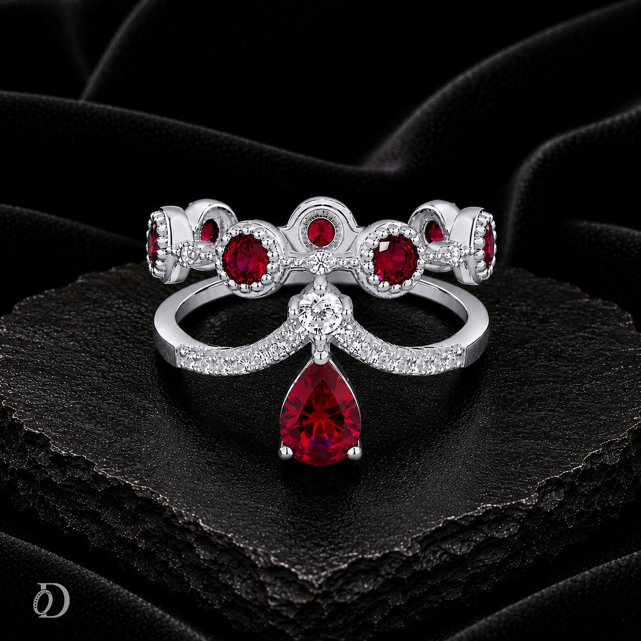 A silver-toned bow-shaped ring set with multiple round and teardrop-cut red gemstones, displayed on a textured black surface against a dark draped fabric background.