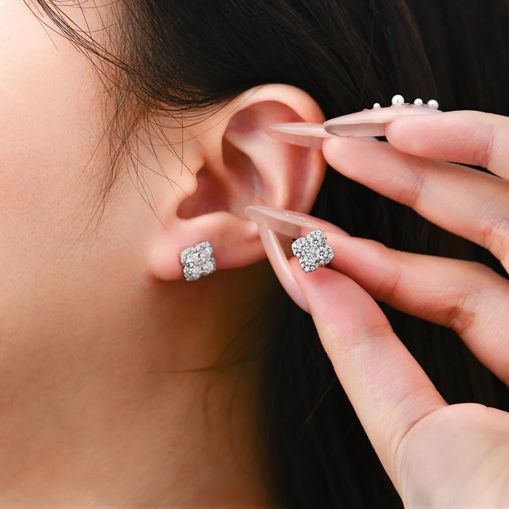 A close-up of a person's ear wearing a small square stud earring, with another matching earring held near the ear by a hand with long clear acrylic nails.
