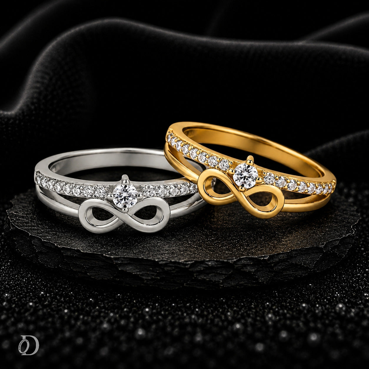 Two infinity-style rings with pavé-set clear stones, one in polished yellow gold and one in polished silver, displayed on a dark textured surface with draped black fabric behind them.
