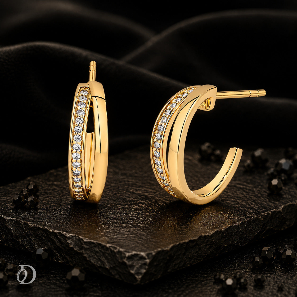 A pair of gold hoop earrings with rows of clear pavé stones, displayed on a dark textured stone slab with black fabric in the background.