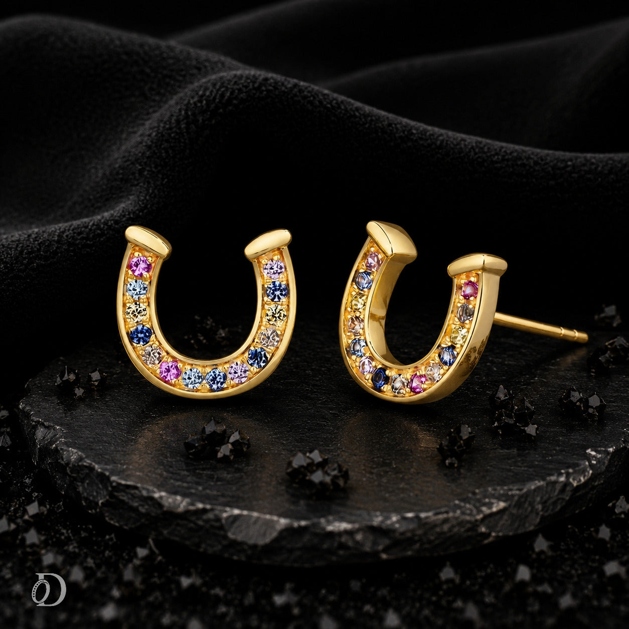 A pair of gold horseshoe stud earrings decorated with small multicolored gemstones, displayed on a dark textured surface with a black fabric backdrop.