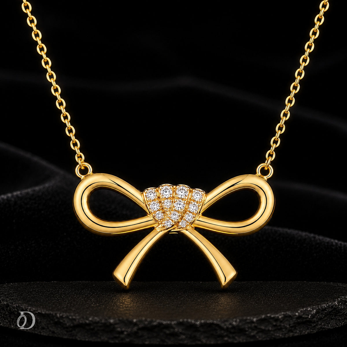 A gold bow pendant necklace with a cluster of small clear stones at the center, displayed on a black satin background.
