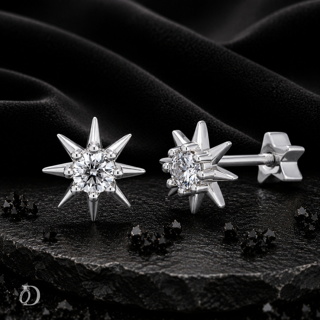 A pair of silver starburst stud earrings with clear gemstones, displayed on a textured black surface with draped black fabric in the background.