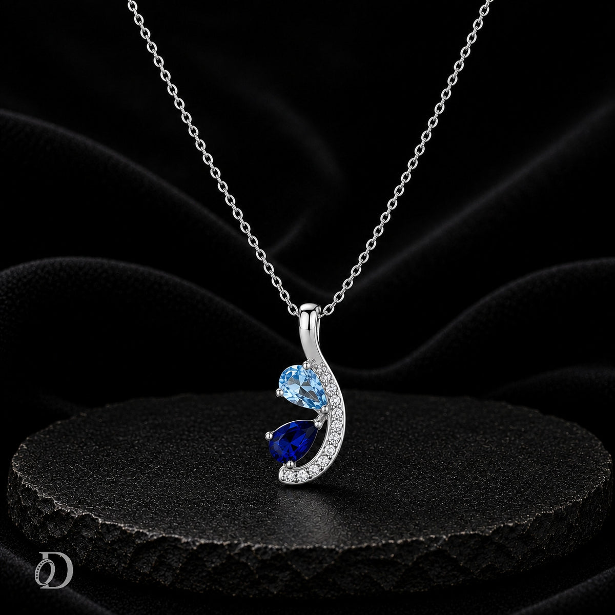 A silver pendant necklace with a curved wave design, featuring a light blue round gemstone, a larger dark blue pear-shaped gemstone, and small clear accent stones, displayed on a black textured surface against a black fabric background.
