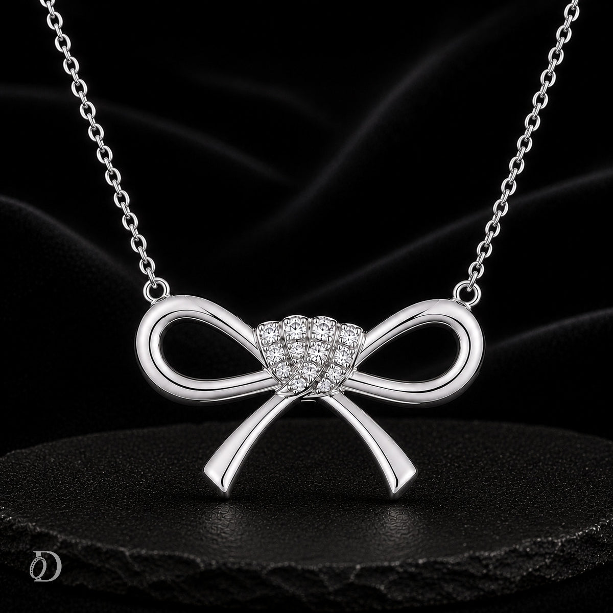 A silver bow pendant necklace with a heart-shaped cluster of small clear stones at the center, displayed on a black satin background.
