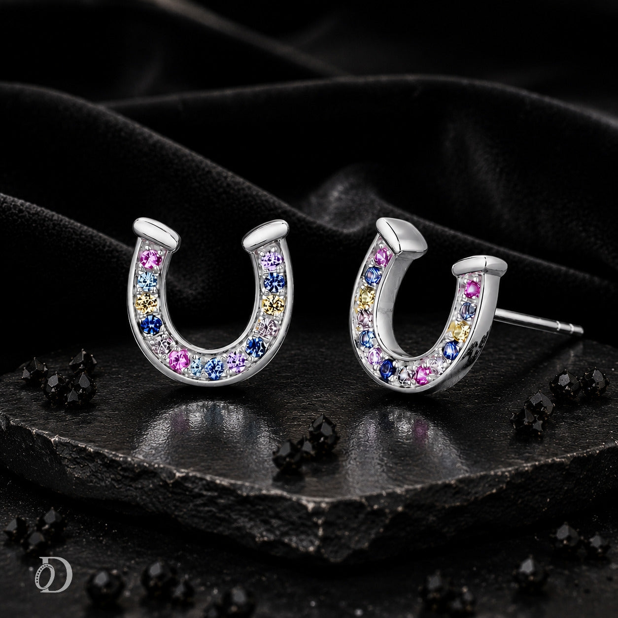 A pair of silver horseshoe stud earrings set with small multicolored gemstones in pink, blue, yellow, and white, displayed on a dark textured stone surface with black fabric in the background.