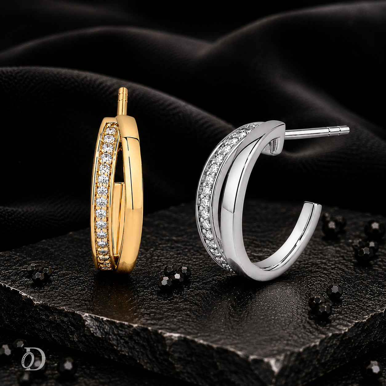 Two small hoop-style earrings displayed on a dark textured surface with draped black fabric behind them. One earring is gold with a row of clear stones, and the other is silver with a row of clear stones.