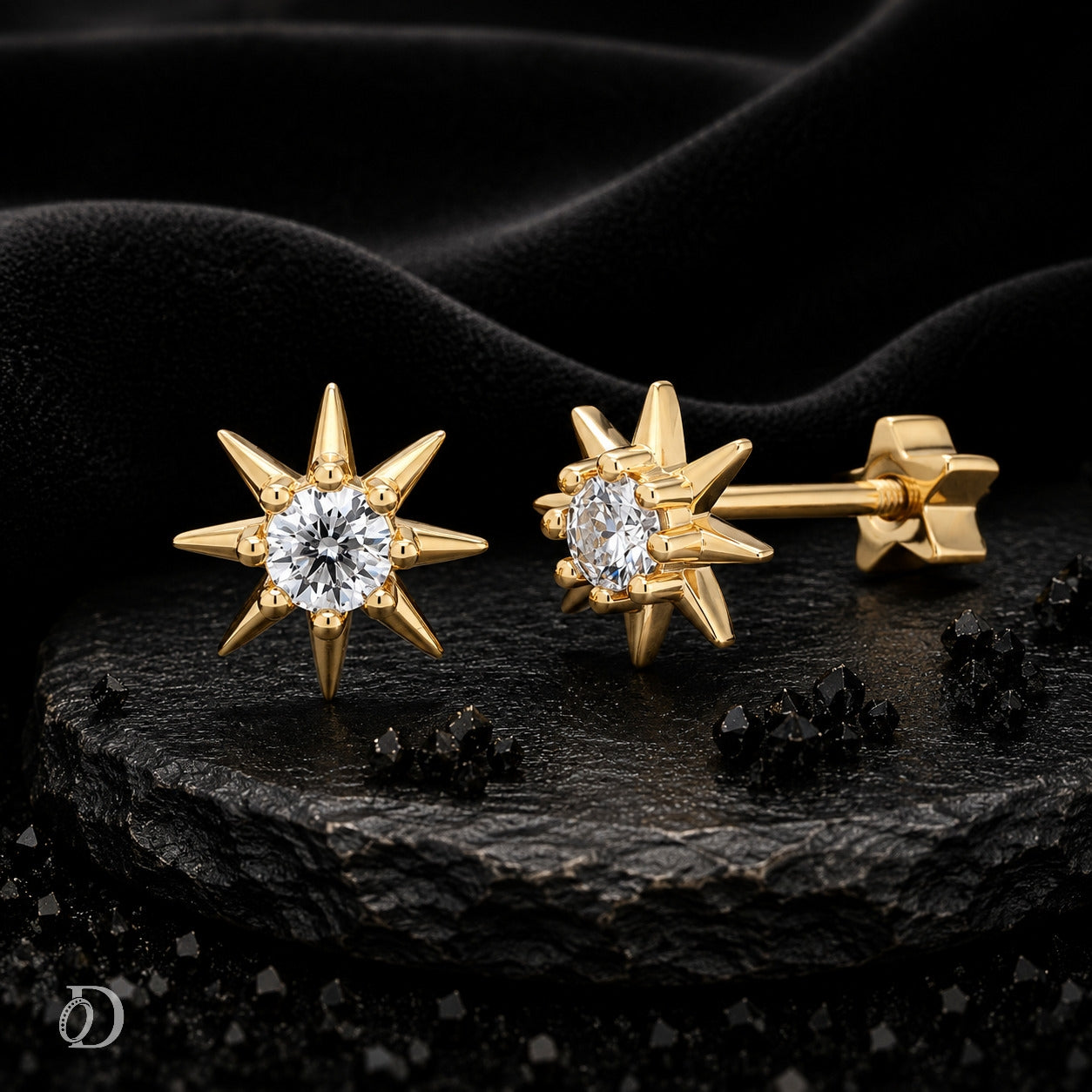 A pair of gold starburst stud earrings with clear round-cut gemstones, displayed on a dark textured stone slab against a black fabric background.