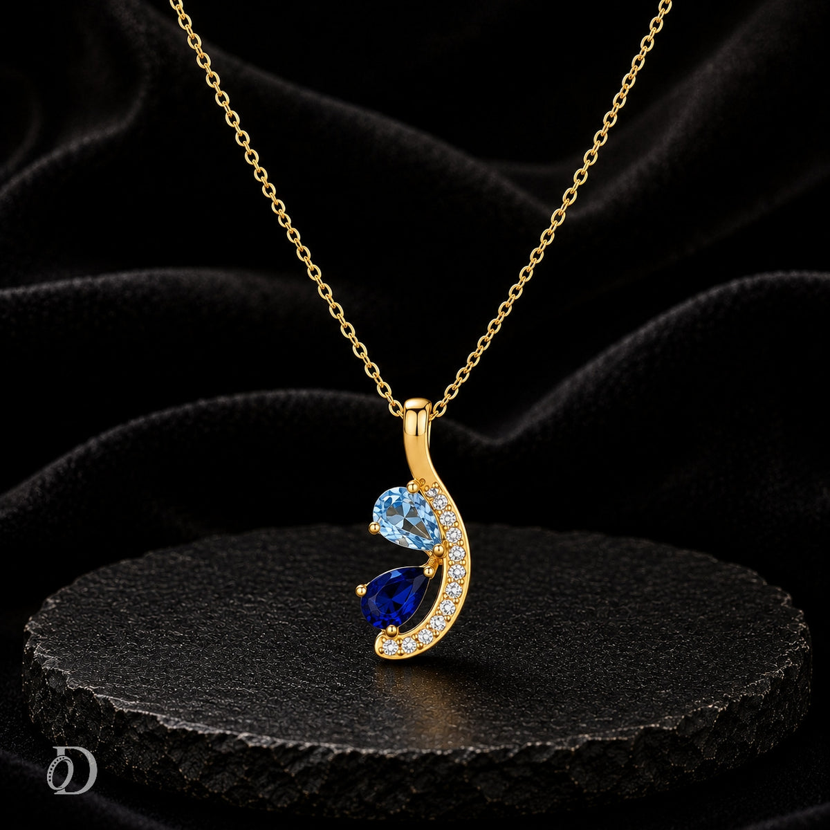 Gold crescent moon pendant necklace with a light blue top and dark blue bottom stone, accented with small clear stones, displayed on a black textured surface against a black fabric background.